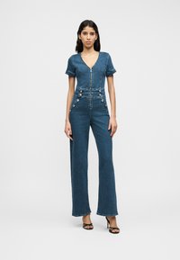 Ολόσωμη φόρμα από τζιν σε μεσαίο μπλε με κοντά μανίκια, λαιμόκοψη σε σχήμα V, φερμουάρ μπροστά και διακοσμητικά κουμπιά στο πλάι. Σχέδιο με φαρδιά μπατζάκια.