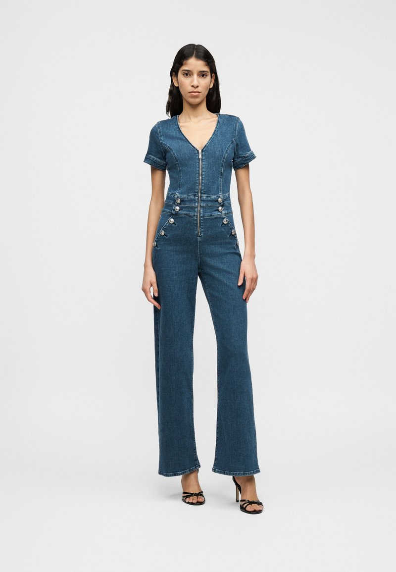 Ολόσωμη φόρμα από τζιν σε μεσαίο μπλε με κοντά μανίκια, λαιμόκοψη σε σχήμα V, φερμουάρ μπροστά και διακοσμητικά κουμπιά στο πλάι. Σχέδιο με φαρδιά μπατζάκια.