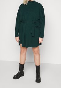Abito verde scuro con maniche lunghe, collo alto e cintura in vita. Abbinato a stivali neri alla caviglia. Tessuto liscio.