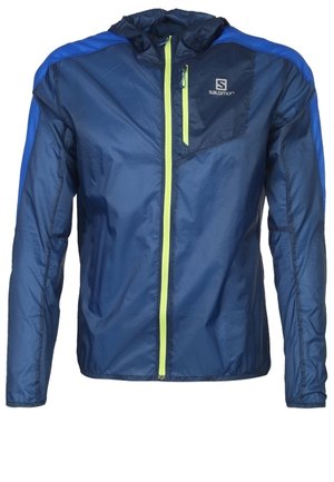 Mand iført en blå lynlåstrøje over en hvid skjorte, sorte ASICS leggings og mørkeblå løbesko med orange detaljer.