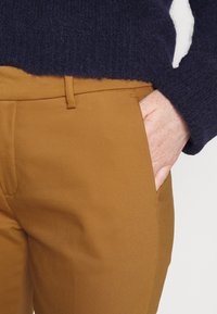 Bruna skräddarsydda byxor med en slät textur, som har en sidoficka och en platt framsida. I kombination med en mörk marinblå stickad tröja.