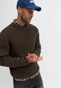 Pullover in maglia grossa marrone con colletto alto, indossato sopra una camicia a quadri rossa e bianca. Le mani sono incrociate in modo informale davanti.