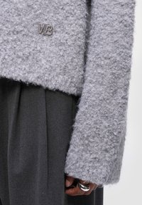 Šedý, texturovaný svetr s chlupatým povrchem a zkráceným střihem, s kovovým logem "WB". V kombinaci s tmavými plisovanými kalhotami.