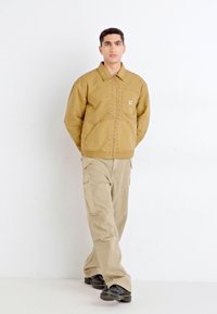 Giacca di cotone marrone con tasca anteriore, abbinata a pantaloni cargo beige. Il modello è in piedi su uno sfondo bianco. Scarpe nere completano il look.