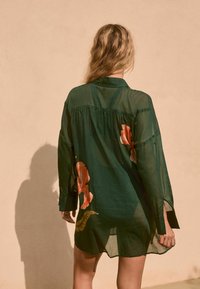 Groene, halfdoorzichtige blouse met lange mouwen; heeft grote oranje bloemmotieven; kraagontwerp; losse pasvorm met afgeronde zoom.