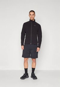 Juodas vilnonis švarkelis su aukštu apykakliu ir užtrauktuku priekyje, derinamas su juodais šortais su keliais kišenėmis ir juodais sportiniais bateliais.