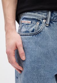 Svetlo modré džíny z denimu s textúrovaným povrchom. So známkou "Calvin Klein" v červenej a bielej farbe na prednej vrecku.