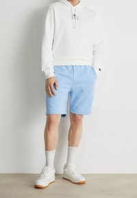 Vit hoodie med en liten grafik, kombinerad med ljusblå shorts. Accessoarer inkluderar vita sneakers med gummisulor och vita strumpor.