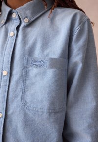 Vaaleansininen farkkukankaasta valmistettu napillinen paita, jossa on päällekirjottu tasku ja rintalogo. Pehmeä materiaali ja klassinen muotoilu.