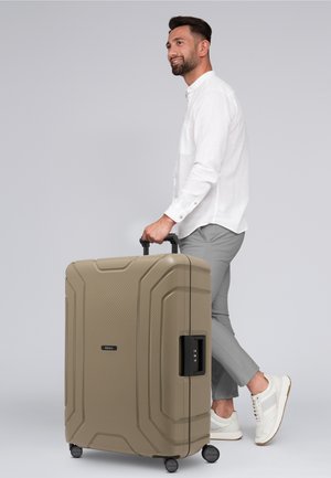 Harde koffer in een gestructureerd beige, met een zwart combinatieslot en vier wielen, vastgehouden door een persoon in lichtgrijze broek en witte shirt.