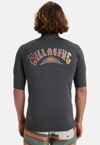 Mörkgrå rash guard med korta ärmar och en färgglad "Billabong"-logotyp med stiliserad text och mönster över ryggen.