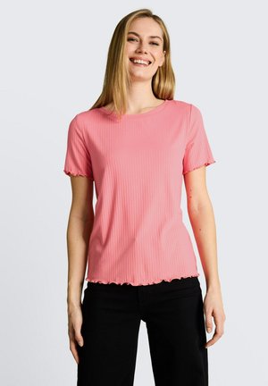 Femme souriante aux cheveux blonds et raides, portant un t-shirt côtelé rose à manches courtes et un pantalon noir, debout devant un fond uni.