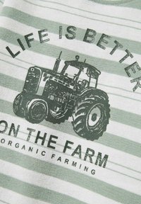 Zielono-biała materiał z paskami przedstawia grafikę traktora z czarnym napisem: "ŻYCIE JEST LEPSZE NA FARMIE", podkreślając rolnictwo ekologiczne.