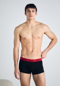 Čierne boxerky s červeným elastickým pásom označeným "HUGO." Materiál vyzerá hladko a je priliehavý. Dizajn je minimalistický a funkčný.
