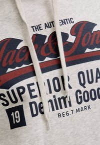 Felpa beige con coulisse crema e testo "The Authentic Jack & Jones Superior Quality Denim Goods 19 Reg.T.Mark" stampato in nero e rosso.