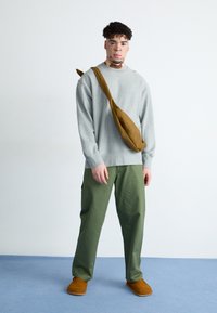 Sudadera gris, pantalones cargo verdes, bolsa cruzada marrón y zapatos marrones de slip-on. El conjunto presenta un ajuste relajado y texturas simples.