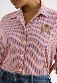 Žena oblečená v košeli s vertikálnymi pruhmi v červenej, modrej a bielej farbe, s vyšívaným erbom na hrudi, a modrých rifliach.