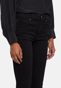 Jeans de mezclilla negros con corte ajustado, que presentan bolsillos frontales, costuras sutiles y una textura suave. Se llevan con una blusa de manga larga oscura.