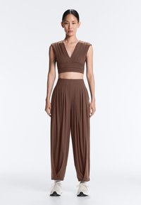 Femme debout face à l’objectif, portant un crop top marron sans manches avec des plis et un pantalon plissé taille haute assorti ainsi que des baskets blanches.