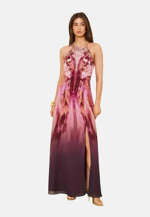 Woman wearing a sleeveless, halter-neck, floor-length purple and pink patterned dress with a high slit and gold strappy heels.