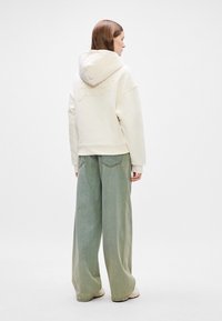 Cremfarbene Kapuzenjacke mit aufgesticktem "Tommy Jeans" auf dem Rücken, kombiniert mit weit geschnittenen grünen Jeanshosen und weißen Turnschuhen, minimaler Hintergrund.