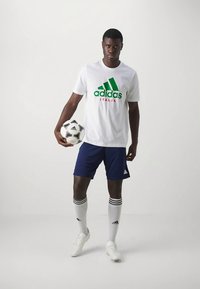 Camiseta blanca de Adidas con logo verde y "ITALIA" en rojo, pantalones cortos azules, calcetas blancas con rayas negras, sosteniendo un balón de fútbol negro y blanco.
