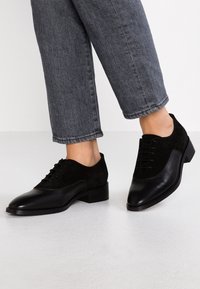 Chaussures en cuir noir et en daim avec un bout pointu, un design à lacets et un petit talon carré. La tige texturée contraste avec le cuir lisse.