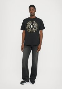 Tricou din bumbac negru cu un logo circular mare de culoare aurie care spune "VERSACE JEANS COUTURE." Purtați-l cu blugi gri închis și adidași negri.