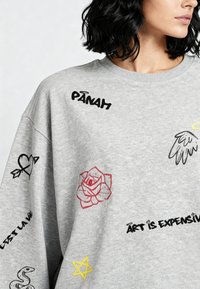 Felpa grigia oversize con vari design ricamati colorati, tra cui cuori, rose, ali e scritte decorative. Texture morbida.