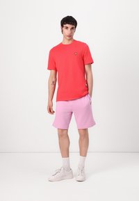 Röd bomulls-t-shirt med en gul logotyp, kombinerad med ljusrosa shorts och vita sneakers, på en helt vit bakgrund.