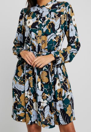 Woman wearing long-sleeve dress with green, mustard, black, white floral pattern, gathered waist, and round neckline against plain background.
