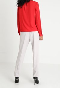 Röd långärmad blus med en slits bak, matchad med ljusgrå skräddarsydda byxor och svarta högklackade skor. Slät tygtextur.
