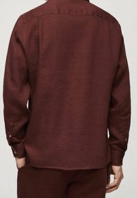 Mann in einem langärmeligen, knöpfbaren, dunkelroten Hemd und dazu passenden Hosen, von hinten vor einem schlichten Hintergrund gezeigt.