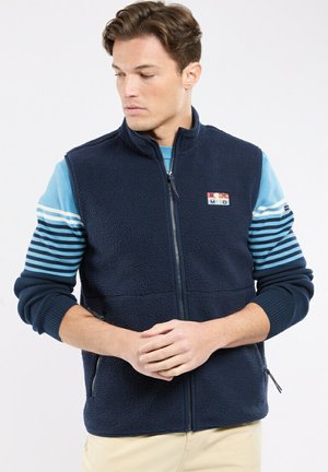Homme portant un gilet polaire bleu marine par-dessus un pull à rayures bleu clair, avec un pantalon beige, regardant vers le bas et les mains jointes devant lui.