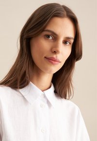 Chemise en lin blanche avec un col classique, dotée d'une fermeture à boutons et d'un tissu légèrement texturé. Le modèle a les cheveux longs et raides de couleur brune.