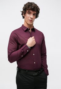 Camisa de manga larga burdeos con cuello abotonado, con botones blancos y textura lisa, usada con pantalones negros y un reloj.