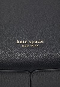 Svart texturerad läderyta med en guld "kate spade New York" logotyp centrerad längst ner. Rektangulär form med subtila sömmar.