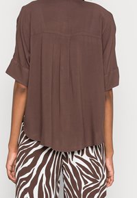 Bruine overhemd met korte mouwen en een relaxte pasvorm, met plooien aan de achterkant en een gladde textuur, gecombineerd met witte broek met een bruin zebra-strepenpatroon.