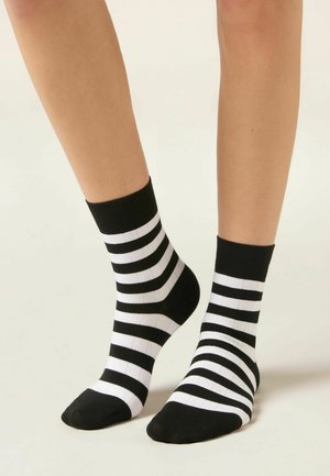 Piedi che indossano calzini alla caviglia a righe orizzontali bianche e nere su uno sfondo chiaro uniforme.