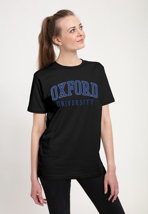 Ung kvinde med brunt hår i hestehale iført sort Oxford University t-shirt, der kigger opad, stående mod en ensfarvet lys baggrund.