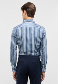 Camicia a maniche lunghe con righe blu in tonalità chiare e scure, dotata di colletto con bottoni e tessuto testurizzato. Abbinata a pantaloni scuri.