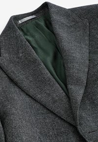 Sacou gri din țesătură tweed cu un material texturat, având o căptușeală din satin verde închis și rever în vârf. Fără hardware sau accente vizibile.