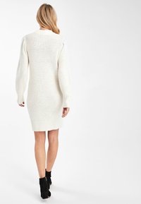 Femme aux cheveux blonds portant une robe pull blanche en maille et des bottines noires, debout de dos face à l'appareil photo.