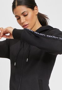 Black zip-up hoodie with adjustable drawstrings, ribbed cuffs, and logo detail along the sleeve. Soft fabric with a smooth texture.