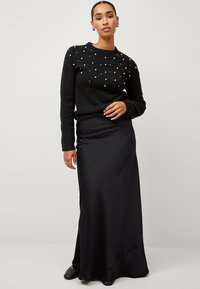 Czarny sweter z perłowymi zdobieniami z jednej strony, zestawiony z elegancką, czarną satynową spódnicą maxi. Całość dopełniają czarne buty.