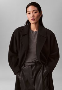 Femme aux cheveux foncés et mouillés porte un manteau noir oversize, un pull gris et un pantalon en cuir noir avec les mains dans les poches devant un fond gris uni.