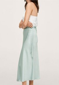 Una lunga gonna in satin verde menta con vita legata e una texture liscia, abbinata a un top bianco senza spalline. Presentazione vista laterale.