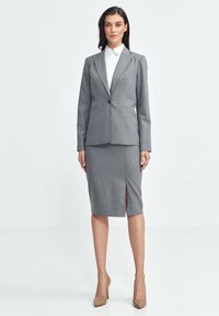 Szary żakiet w prążki z pasującą spódnicą do kolan. Żakiet ma jeden guzik, klapki i rozcięcie w spódnicy.