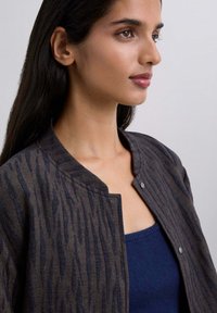 Chaqueta de patrón oscuro con una sutil textura a rayas, que presenta un escote redondo y cierre a presión, superpuesta a una camiseta azul oscura.