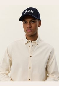 Navy cap with "LES DEUX" embroidered on the front, paired with a cream-colored button-up shirt featuring a textured fabric and visible buttons.